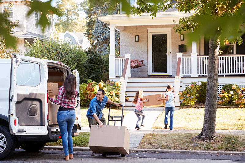 A family is moving into a new house; a man wheels a box on a dolly, a woman loads a van, and two children carry boxes up the walkway. The house has a porch and is surrounded by trees and bushes.