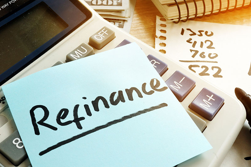 Refinance 1 A sticky note with the word Refinance is placed on a calculator. In the background, there are scattered papers with handwritten numbers and a spiral notebook.