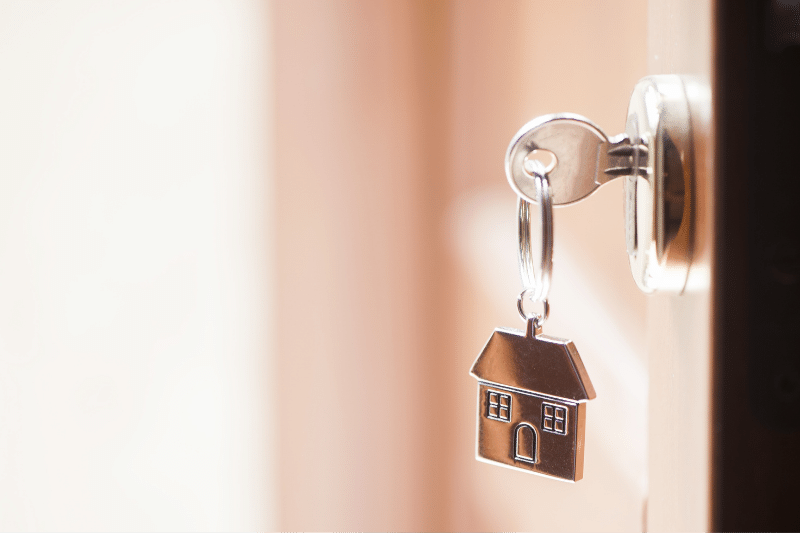 A silver key with a house-shaped keychain is inserted into a door lock, with soft natural light shining from the left side of the image.
