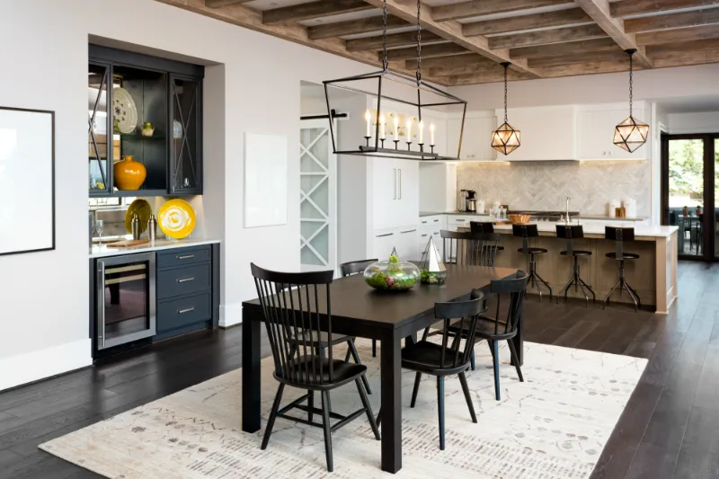 Modern open-concept kitchen and dining area with dark wood floors, black dining table and chairs, white cabinets, bar stools at a kitchen island, pendant lighting, and exposed wood-beam ceiling.