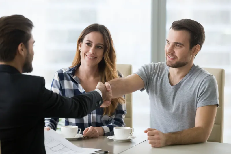A man and woman sit at a table with coffee cups, smiling as the man shakes hands with another man in formal attire, suggesting a successful meeting or agreement.