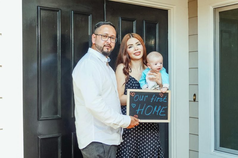 Young family with baby holding a sign Our First Home at door 1774371664